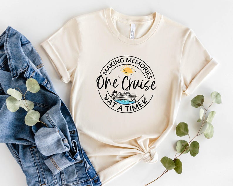 Peut inclure: T-shirt cr&egrave;me &agrave; manches retrouss&eacute;es avec un motif circulaire indiquant "Making Memories One Cruise at a Time". Le motif comprend un bateau de croisi&egrave;re, le soleil, des oiseaux et une ancre. Une veste en jean et de la verdure sont &agrave; c&ocirc;t&eacute; du t-shirt.