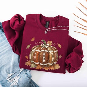 May include: A maroon crewneck sweatshirt featuring a graphic of a brown football-textured pumpkin with white laces, surrounded by scattered autumn leaves. A silver-toned toggle necklace rests on the neckline, paired with distressed blue jeans.