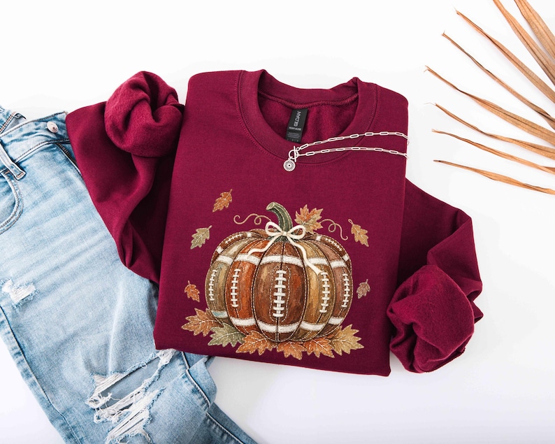 May include: A maroon crewneck sweatshirt featuring a graphic of a brown football-textured pumpkin with white laces, surrounded by scattered autumn leaves. A silver-toned toggle necklace rests on the neckline, paired with distressed blue jeans.
