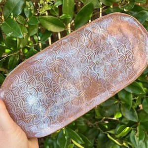 May include: A handmade, oblong ceramic serving tray with a textured, repeating fan pattern. The tray has a brown base color with iridescent blue and white accents. The edges are slightly raised, and the overall shape is oval.