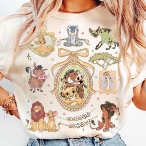 May include: Cream-colored t-shirt featuring cartoon illustrations of characters from The Lion King. The design includes Simba, Timon, Pumbaa, and Scar, along with the phrase "Hakuna Matata". The shirt has a crew neck and short sleeves.