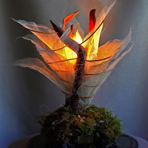Op de afbeelding: Een unieke decoratieve lamp in de vorm van een boom, met een centrale stam en takken versierd met doorschijnende, bladachtige structuren. De bladeren worden verlicht met warm oranje en geel licht, waardoor een zachte gloed ontstaat. De basis is bedekt met groen mos.