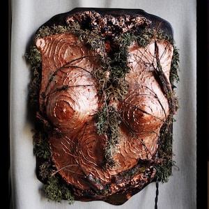 Peut inclure: Sculpture murale cuivrée avec une surface texturée, représentant un torse. La sculpture est ornée de mousse verte et de fines branches sombres, créant une esthétique naturelle et organique. Les bords sont brun foncé.