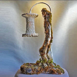 May include: A unique, handcrafted lamp featuring a woven lampshade suspended from a curved support. The base is a natural, moss-covered branch, creating an organic and artistic design. The lamp's design incorporates natural elements.