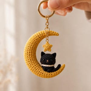 May include: A crochet keychain featuring a yellow crescent moon, a small yellow star, and a black cat with a white collar. The cat sits on the moon, which is attached to a gold-toned metal ring by a short chain. The entire piece is crafted from yarn.