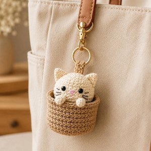 May include: A crochet keychain featuring a small cream-colored cat with black eyes and a pink nose, sitting inside a tan woven basket. The cat figure is attached to a gold-toned metal lobster clasp and ring, hanging from a beige tote bag.