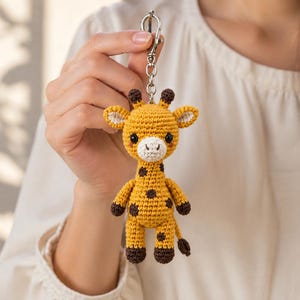 May include: A crochet giraffe keychain featuring a yellow body with dark brown spots, a white muzzle, and small dark brown horns. The stuffed animal is attached to a silver-toned metal lobster clasp and a short chain, showcasing detailed yarn stitching.