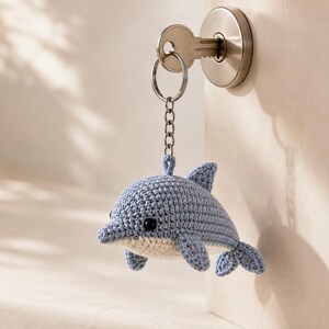 May include: A light blue and white crocheted dolphin keychain featuring a small metal ring and chain. The dolphin has a rounded body, a dorsal fin, side fins, and a tail, with two small black bead eyes attached to the textured yarn surface.