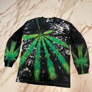 May include: Long-sleeved tie-dye shirt with a large green cannabis leaf design on the front and sleeves. The shirt features a black and white background with a green leaf pattern. The sleeves are long and the shirt is made of a soft material.