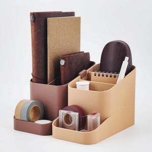 May include: Two desk organizers in shades of brown and tan. One holds notebooks and a journal, the other has a small notepad, pen, and tape. The organizers are made of a smooth, matte material and are designed to hold office supplies.
