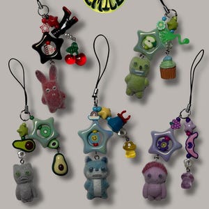 May include: A selection of quirky phone charms featuring diverse designs. Charms include a star, a bunny, a bear, a cupcake, a mushroom, and a pair of legs. The charms are in various colours, including red, green, blue, and purple. The charms are attached to black cords.