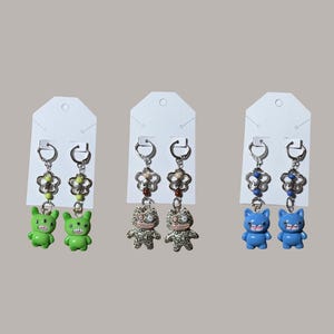 May include: Three pairs of dangle earrings. Each pair features a different design: green monster figures, patterned cat-like figures, and blue monster figures. The earrings have silver-colored hoops and flower-shaped accents with colorful beads.