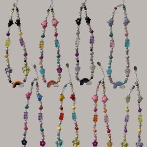 Könnte beinhalten: Eine Sammlung von Halsketten mit bunten Perlen, darunter Sterne, Blumen und Gummibärchen. Jede Halskette hat einen Regenbogen-Anhänger und eine silberne Kette.