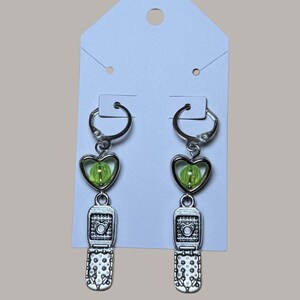 May include: Silver-tone earrings with a heart-shaped green bead and a miniature flip phone charm. The earrings hang from a silver hoop and are attached to a white earring display card.