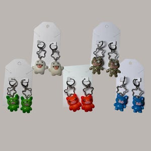 May include: A collection of colorful, quirky earrings featuring various monster-like characters. Each pair is attached to a white card. The earrings are in shades of green, orange, blue, white, and leopard print, with star-shaped accents and silver hoops.