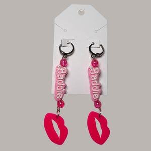 Puede incluir: Un par de pendientes colgantes con aros negros. Cada pendiente presenta un dije rosa con la palabra "Baddie" en blanco, cuentas rosas y un dije en forma de labio rosa brillante. Los pendientes cuelgan de una tarjeta blanca.
