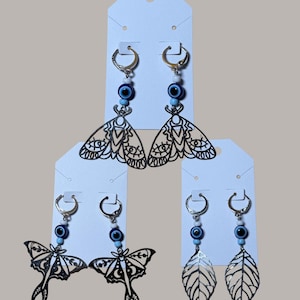 May include: Earrings with silver-tone hoops, blue beads, and decorative charms. The charms include two moth designs and a leaf design. The earrings are displayed on white earring cards against a neutral background.