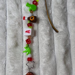 Puede incluir: Un marcapáginas de cuentas con una parte superior de metal dorado. Las cuentas incluyen una botella verde, una botella marrón, un cubo blanco con texto rojo, dados rojos, una hoja verde, una cuenta en forma de estrella y una hamburguesa.