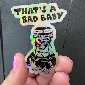 Pode incluir: Um autocolante holográfico com uma ilustração gráfica preta de uma criatura a usar óculos de proteção e uma fita para a cabeça, com o texto THAT'S A BAD BABY impresso a preto por cima. O autocolante tem um acabamento refletor e iridescente.