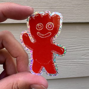 May include: A red, Sour Patch Kids-inspired sticker in the shape of a smiling gummy bear waving. The sticker has a glittery, iridescent border and is held up by a hand.