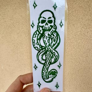 May include: A green and white sticker featuring a skull with a snake wrapped around it. The design includes diamond shapes around the central image. The sticker is in a clear plastic sleeve.