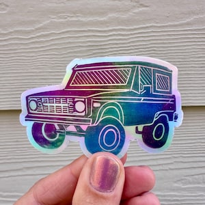 May include: A holographic sticker of a vintage SUV. The sticker has a rainbow sheen with purple, blue, and green hues. The vehicle is outlined in silver, with details like the headlights, grill, and wheels visible.