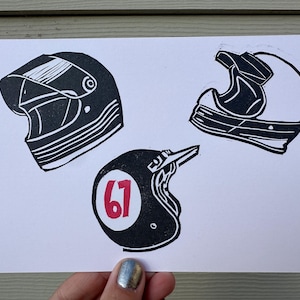 May include: A white card featuring three black motorcycle helmet illustrations. One helmet has a visor, another is open-faced, and the third has the number "67" in a red circle. The artwork is a linocut style.