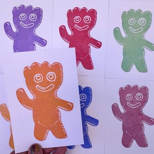 May include: A collection of colorful, hand-printed art cards featuring a smiling, cartoon-like character. The cards showcase the character in various colors, including purple, red, green, orange, blue, and pink, all printed on white cardstock.