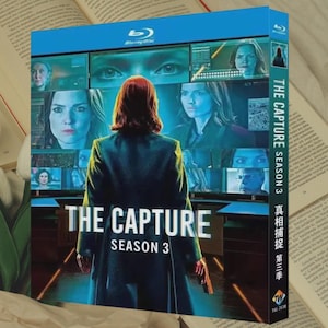 Puede incluir: Caja de disco Blu-ray de "The Capture Temporada 3". La portada presenta a una mujer pelirroja, de espaldas, frente a múltiples pantallas. El título está en letras blancas grandes. La caja es azul y blanca.