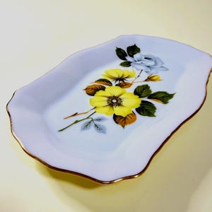 May include: A pale blue ceramic dish with a gold trim and a wavy edge. The dish features a floral design with yellow and blue flowers, green leaves, and brown accents. The dish is rectangular in shape.