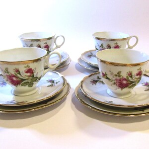 May include: Four white teacups and saucers with gold edging and floral rose patterns. The teacups have handles and a rounded shape. The saucers are scalloped and trimmed with gold. The roses are pink and green.