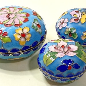 May include: Three blue cloisonné trinket boxes with floral and butterfly designs. The boxes have gold trim and colorful enamel flowers in pink, yellow, and red. The largest box is oval-shaped. The measurements are in centimetres.