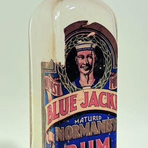 May include: Vintage clear glass bottle of "Blue Jack" matured Normanby Rum. The label features a sailor illustration and text in red and blue. The bottle has a cork stopper.