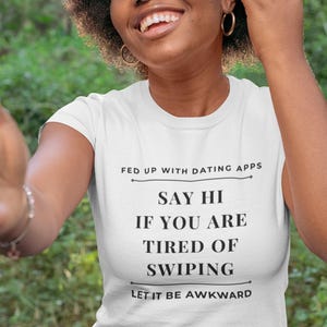 May include: White t-shirt with the text "FED UP WITH DATING APPS, SAY HI IF YOU ARE TIRED OF SWIPING, LET IT BE AWKWARD" in black. The shirt is worn by a person with dark curly hair and gold hoop earrings.