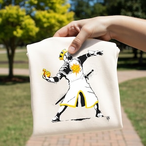 May include: A cream-colored t-shirt featuring a graphic print of a knight wearing a crown and armor with a sun emblem on the chest. The knight is depicted in a dynamic pose, holding a golden orb in one hand, with black and yellow accents.
