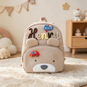 Puede incluir: Mochila de pana beige con orejas en forma de oso y diseño de cara de oso. El nombre "Henry" está bordado en la parte delantera, junto con un coche rojo y un avión azul. La mochila tiene un asa superior y un bolsillo frontal con cremallera.