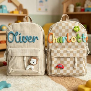 以下が含まれることがあります： ベージュ色のバックパック2つ。1つは、青い糸で「Oliver」という名前とライオンのアップリケが付いたコーデュロイです。もう1つは、カラフルな糸で「Charlott」、花、赤い車のアップリケが付いたチェック柄です。どちらもフロントポケットが付いています。
