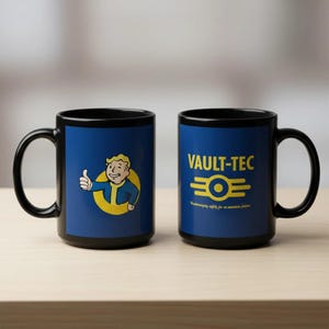 May include: Two black ceramic mugs with blue panels featuring Vault-Tec designs. One mug displays the Vault Boy giving a thumbs up, the other has the Vault-Tec logo and text. The mugs are on a light brown surface.