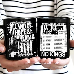 Puede incluir: Dos tazas de cerámica negra con el texto "Springsteen & E Street Land of Hope & Dreams American Tour". Una taza tiene un gráfico de un músico tocando la guitarra. La otra taza enumera las fechas y lugares de la gira. La frase "No Kings" está impresa en la segunda taza.