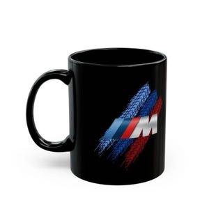 May include: A black ceramic mug featuring a graphic of the letter M in a metallic silver finish, accompanied by diagonal stripes in blue, red, and white. The design is overlaid on a textured pattern resembling tyre tracks in blue and red.