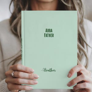 May include: A light green journal with the words "ABBA FATHER" and the name "Heather" printed on the cover. The spine reads "PRAYER JOURNAL". The journal is held by a person.