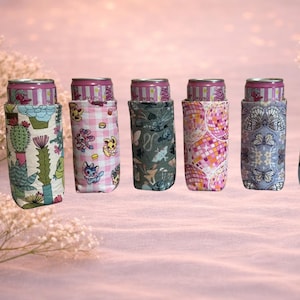 May include: A collection of seven beverage can coolers, each with a unique patterned fabric design. The patterns include cacti, cartoon characters, and butterflies. The cans are partially visible, adding a pop of color to the display.