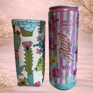 Op de afbeelding: Een stoffen drankhoes met een cactusprint naast een energiedrankblikje met suikersmaak. Het blikje is roze en paars met het woord "Energy" en "Cotton Candy" erop gedrukt. Het blikje is 355 ml.