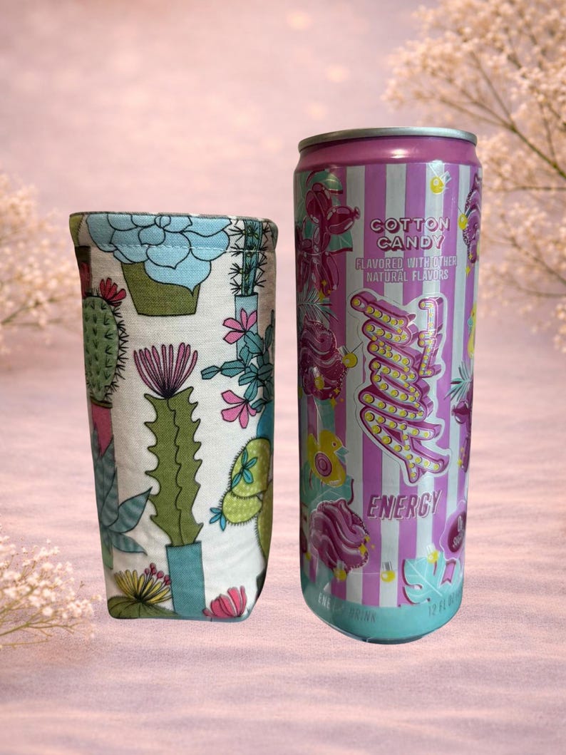 Op de afbeelding: Een stoffen drankhoes met een cactusprint naast een energiedrankblikje met suikersmaak. Het blikje is roze en paars met het woord "Energy" en "Cotton Candy" erop gedrukt. Het blikje is 355 ml.