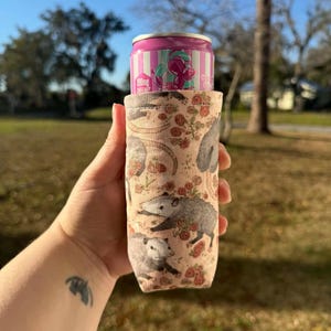 May include: A beige can cooler featuring a pattern of opossums and red roses. A pink and white striped can with text is inside the cooler. The cooler is held in a hand against a background of green grass and trees.