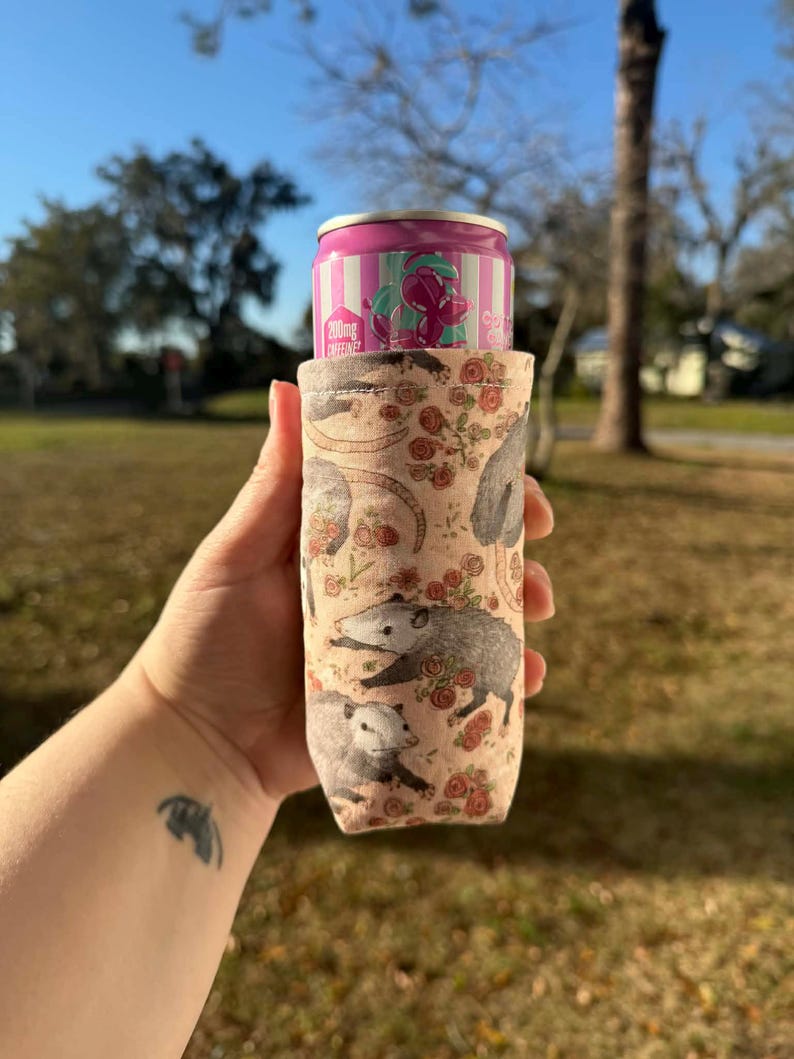 May include: A beige can cooler featuring a pattern of opossums and red roses. A pink and white striped can with text is inside the cooler. The cooler is held in a hand against a background of green grass and trees.
