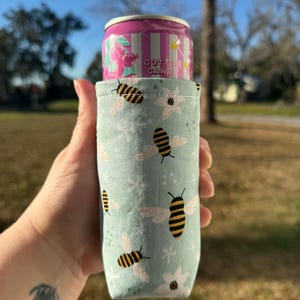 May include: A light blue can cooler with a pattern of bees and flowers, holding a pink and white striped can labeled "Cotton Candy". The cooler is made of fabric and is designed to keep beverages cold.
