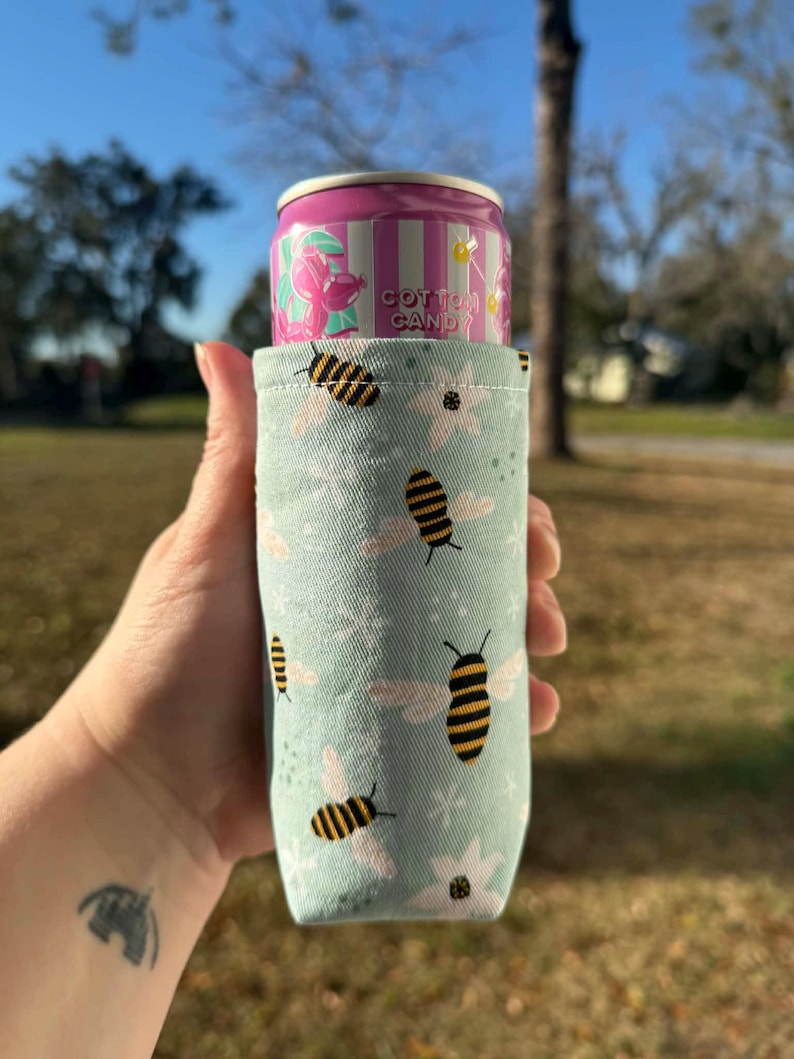 May include: A light blue can cooler with a pattern of bees and flowers, holding a pink and white striped can labeled "Cotton Candy". The cooler is made of fabric and is designed to keep beverages cold.