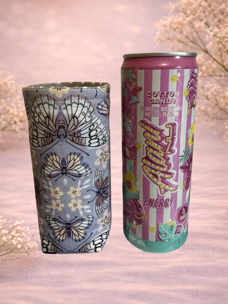 Op de afbeelding: Een stoffen drankhoes met een vlinder- en bloemenpatroon naast een Cotton Candy-gearomatiseerde energiedrankblik. Het blik is roze en wit gestreept met het woord "Energy" in een neonstijl.