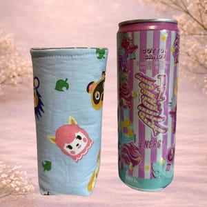 Op de afbeelding: Een lichtblauwe drankhoes met stripfiguren en bladeren. Daarnaast staat een roze en wit gestreepte energiedrankblik met de woorden "Cotton Candy" en "Energy".